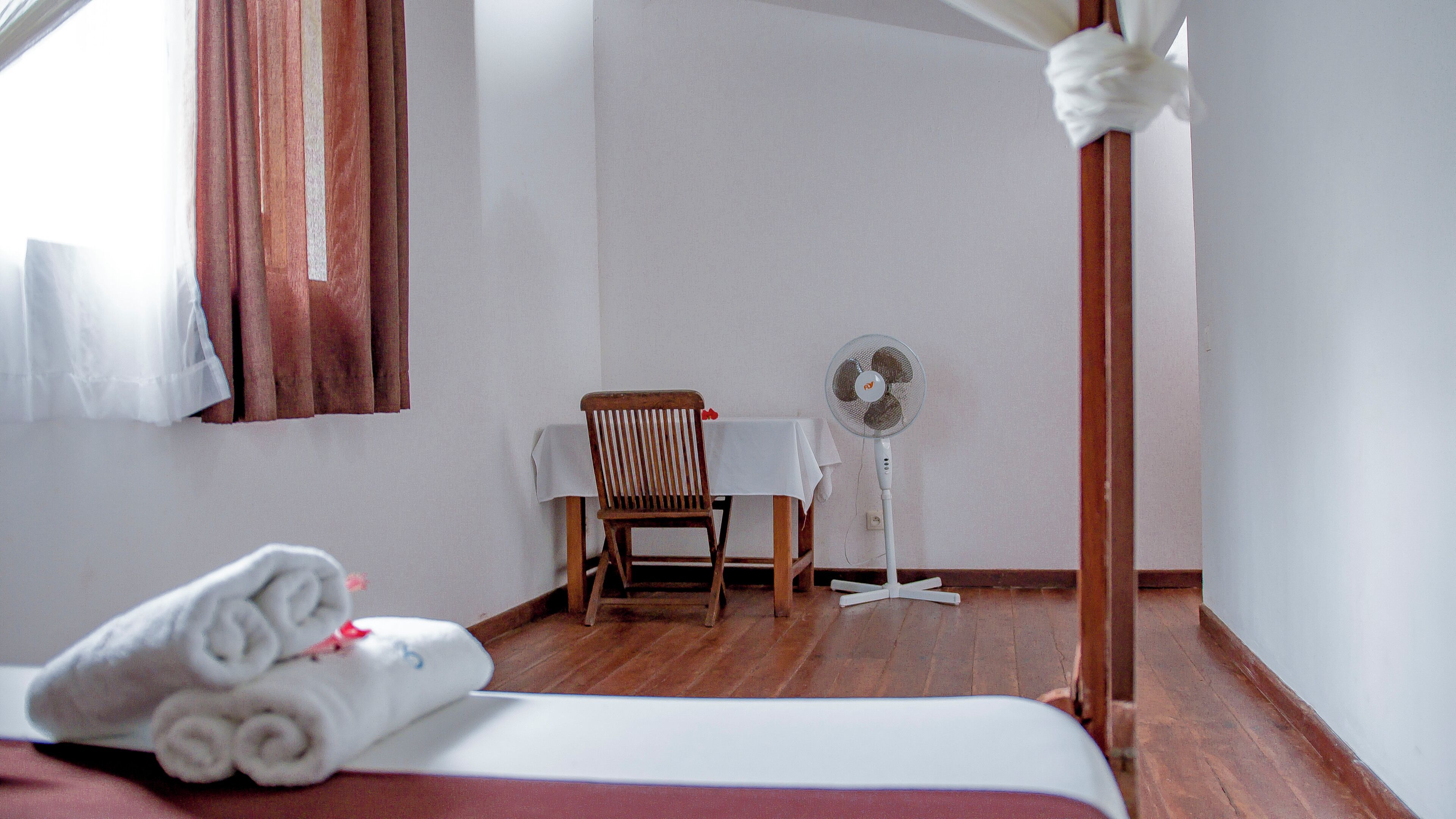 A hotel room with a bed, a chair, a table, and a standing fan.