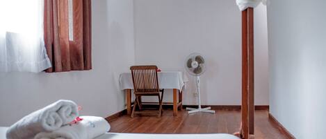 A hotel room with a bed, a chair, a table, and a standing fan.