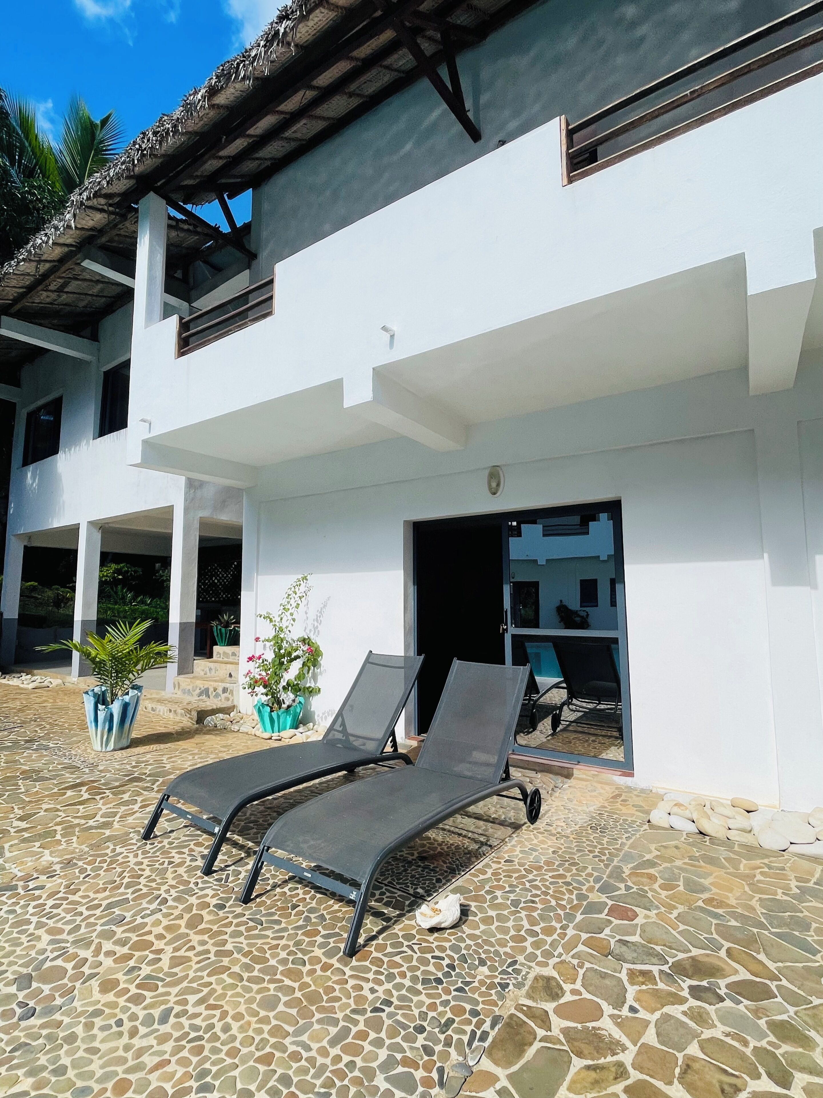 Un edificio con fachada blanca, un patio cubierto con dos sillones de descanso y piso de baldosas de piedra.