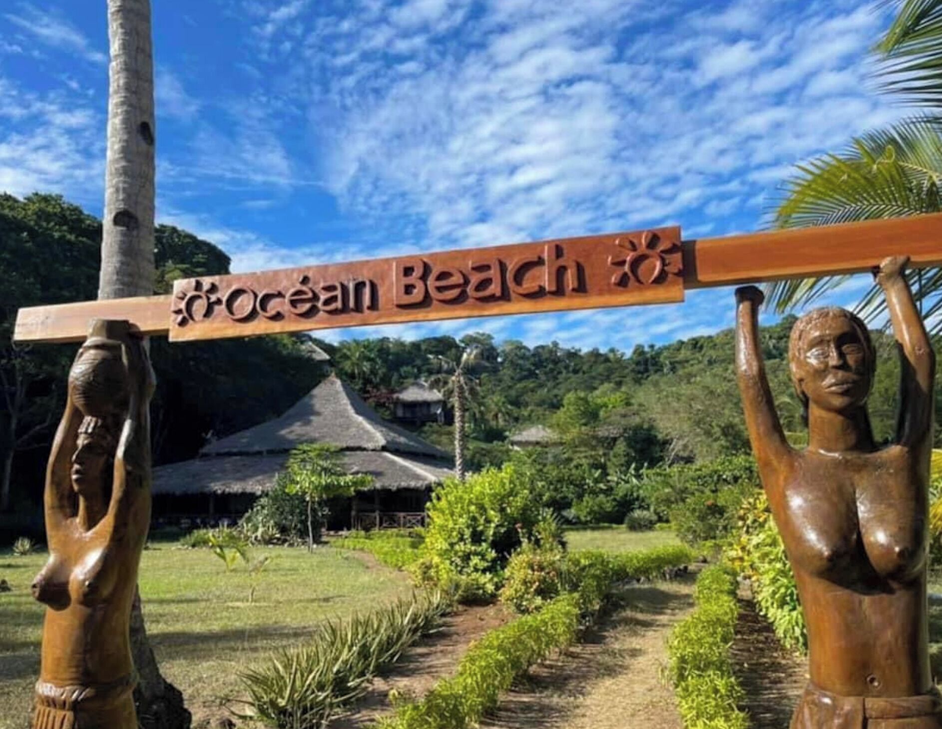 A wooden sign with 'Ocean Beach' written on it, supported by two wooden figures, with a thatched-roof structure and greenery in the background.