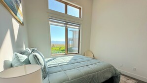 Ein Schlafzimmer mit einem großen Bett, einem Nachttisch, einer Lampe, einem Sessel und einem Fenster mit Blick ins Freie.