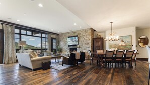 Amplia sala de estar con chimenea, comedor con candelabro y grandes ventanales con vistas al exterior.