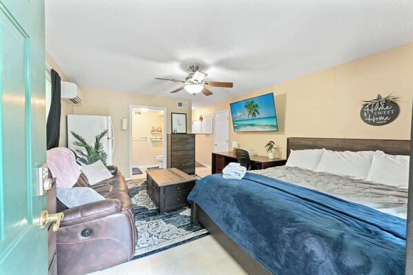 A hotel room with a bed, a desk, a chair, a ceiling fan, a painting, and a wall sign.
