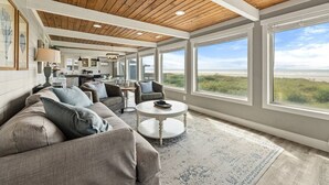 Un salón espacioso con un ventanal grande que da a una playa, una zona de estar acogedora y una cocina al fondo.