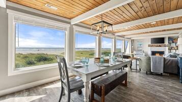 A spacious room with a large window overlooking a beach, a dining area with a wooden table and chairs, and a cozy seating area with a fireplace.
