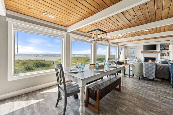 A spacious room with a large window overlooking a beach, a dining area with a wooden table and chairs, and a cozy seating area with a fireplace. - Tide Pool Town Home, Entire House, Amazing Views! (Moclips)