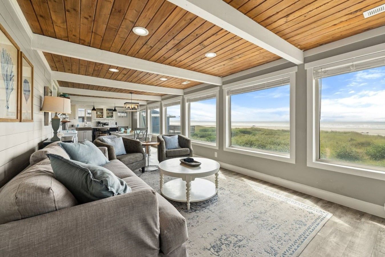 A spacious living room with a large window offering a view of the beach, a cozy seating area with a coffee table, and a kitchen in the background.
