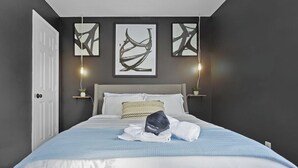 Een net opgemaakt bed met een blauw-witte spreideken, twee wandlampen en abstracte kunst aan de muur.
