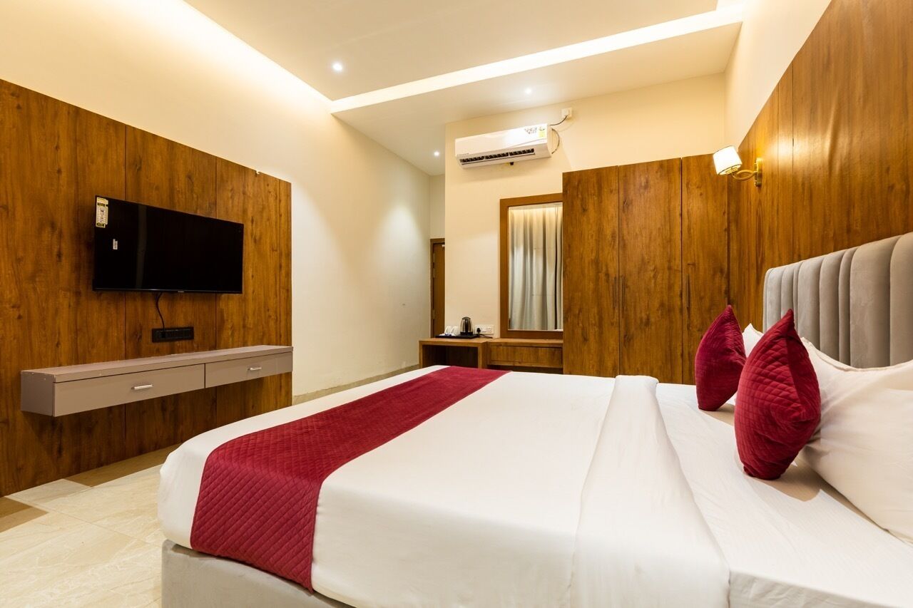 A hotel room with a wooden wall panel, a flat-screen TV mounted above a wooden dresser, a bed with white and red bedding, and a large mirror.