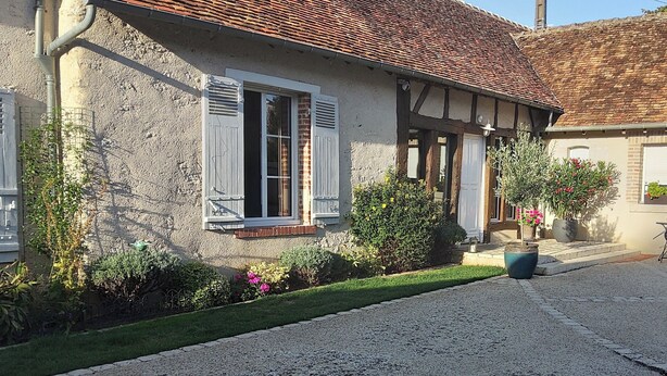 Une maison traditionnelle à colombages, avec une allée gravillonnée, un petit patio doté d’une table et de chaises, et des plantes en pot.