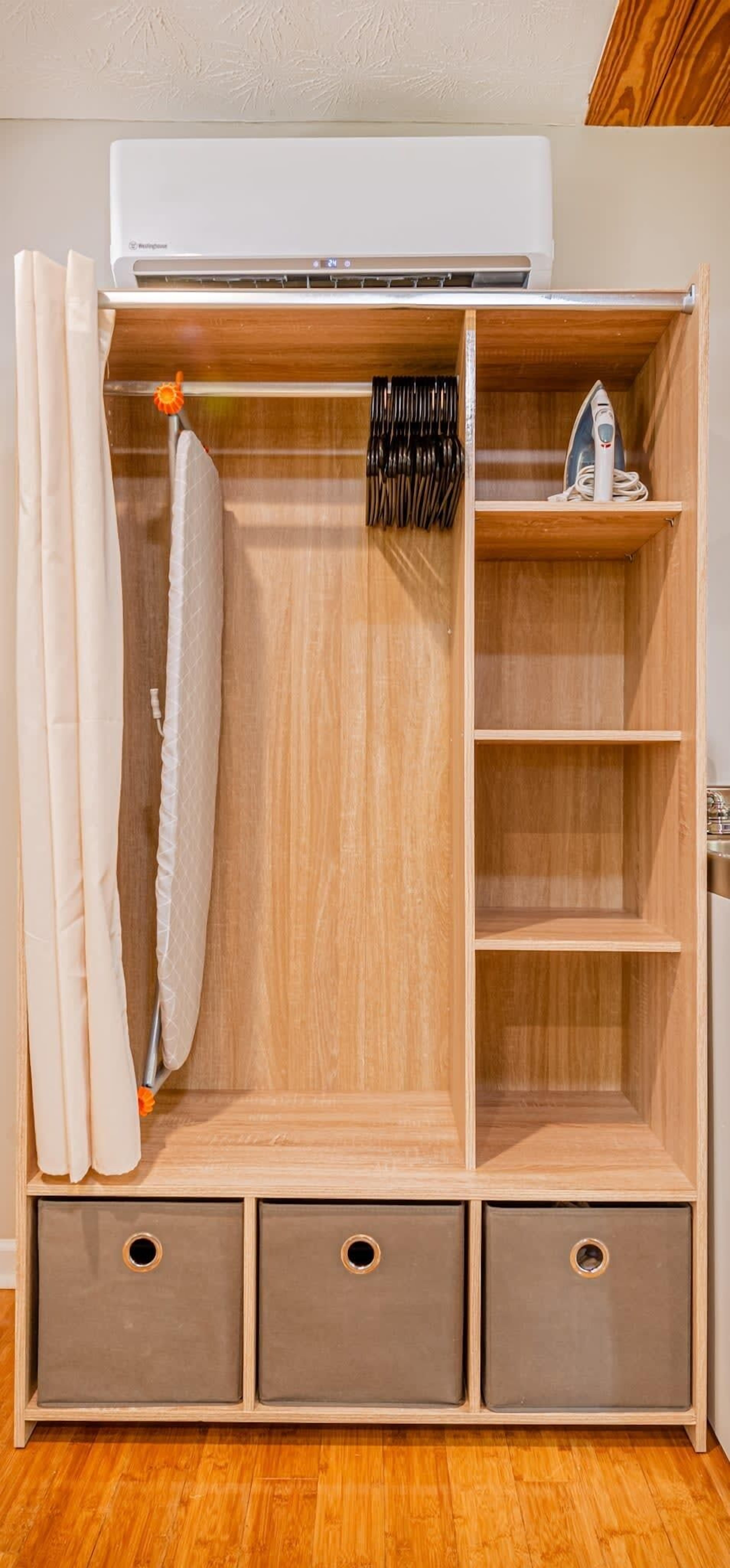 Une armoire en bois équipée d’un climatiseur intégré, d’une tringle à vêtements et d’une étagère avec un fer à repasser.