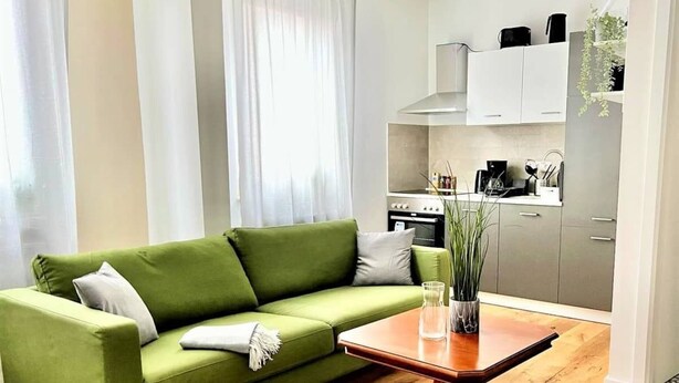 Een groene bank met een witte plaid, een houten salontafel en een keukenhoek met kasten en een plant.