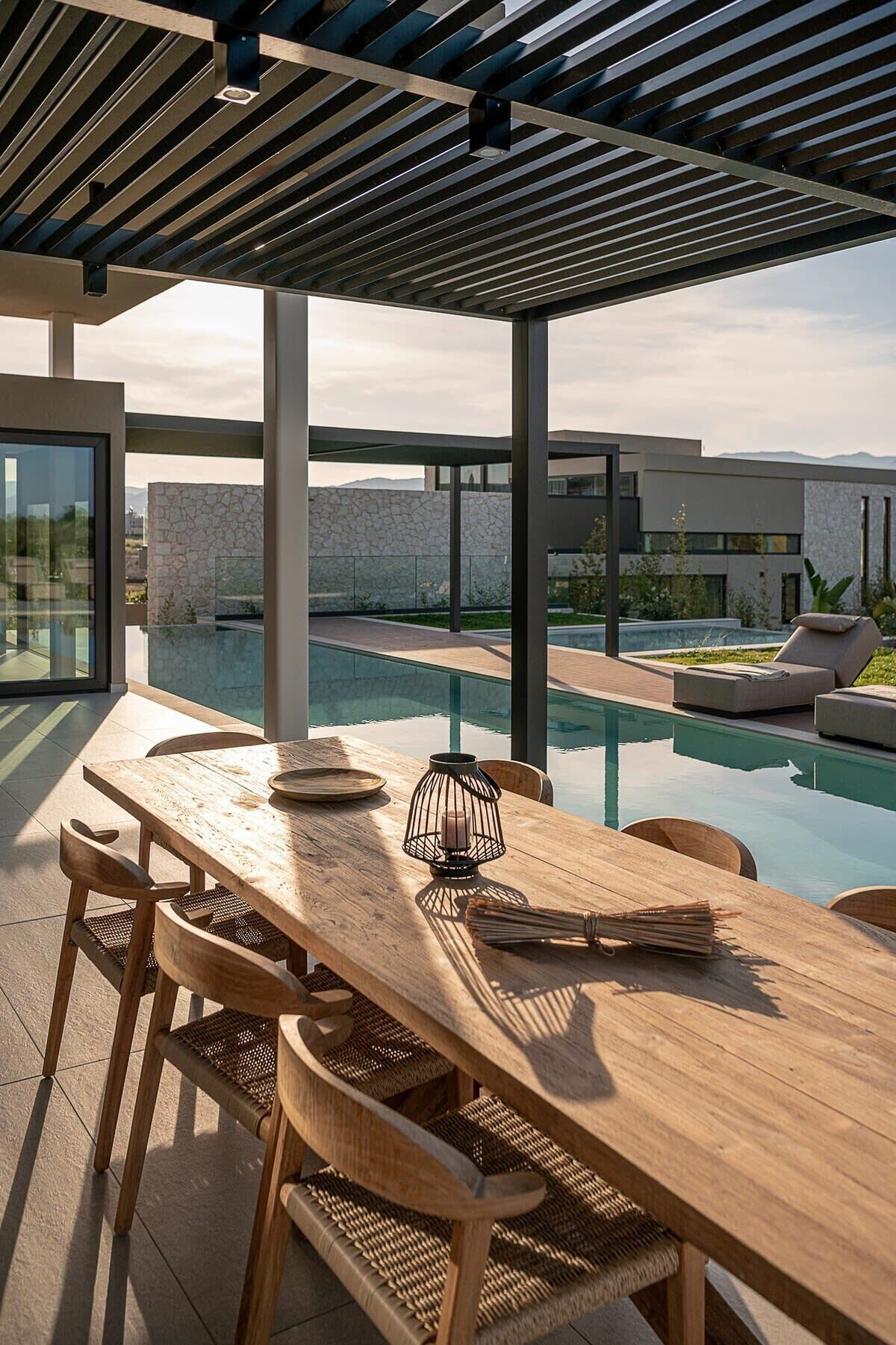 Une table à manger en bois, avec des chaises, une bougie et des journaux, sur une terrasse donnant sur une piscine et un bâtiment moderne.