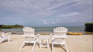 A beach with two white Adirondack chairs and a small table, a yellow boat, and a distant view of the ocean. - Beautiful Waterfront Villa 133 Anglers Reef with On-Site Boat Slip  (Islamorada)