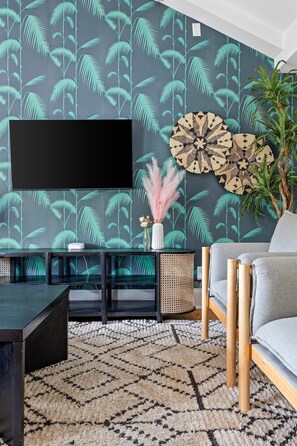 Una sala de estar moderna con un televisor de pantalla plana grande montado en una pared con papel tapiz con estampado de hojas tropicales. Hay una mesa de centro redonda tejida, un sofá gris y una alfombra estampada en el suelo.