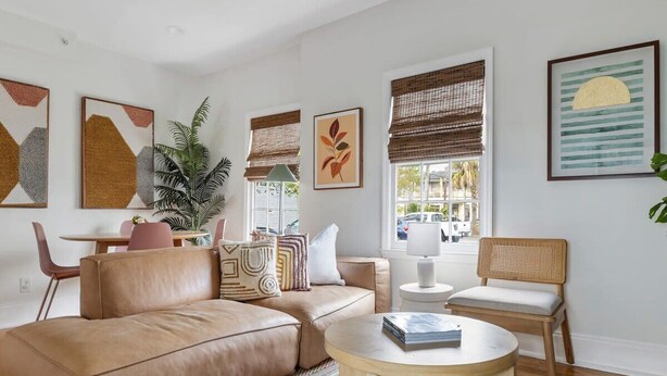 Ein modernes Wohnzimmer mit einem Ledersofa, einem runden Couchtisch und einem Korbstuhl. An den Wänden befinden sich gerahmte Kunstwerke und ein großes Fenster mit Jalousien.