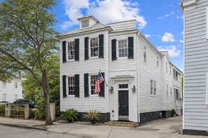A two-story white building with black shutters, a flag, and a small porch with plants.