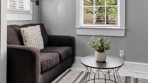 Een woonkamer met een grijze bank, een ronde tafel met een plant, een staande lamp en een raam met zonwering.