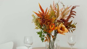 Ein runder Esstisch mit weißen Stühlen, eine Vase mit Herbstblumen und leere Weingläser.