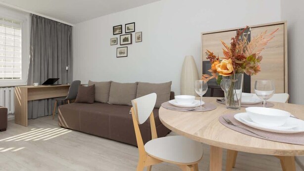 Ein modernes Wohnzimmer mit Sofa, Esstisch und Stühlen. Es gibt ein Fenster mit Vorhängen, eine Heizung und ein an der Wand befestigtes Regal mit Bildern.