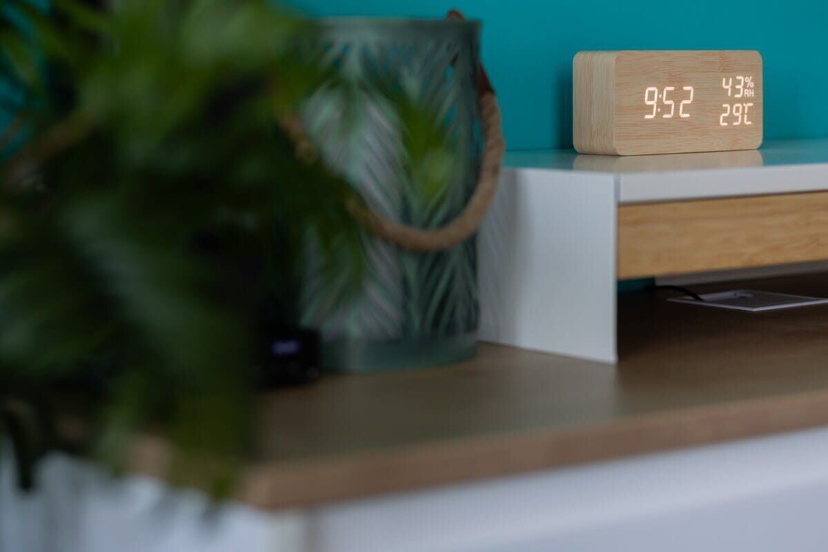 Un reloj digital de madera que muestra las 9:52, una planta y una pared azul verdoso al fondo.
