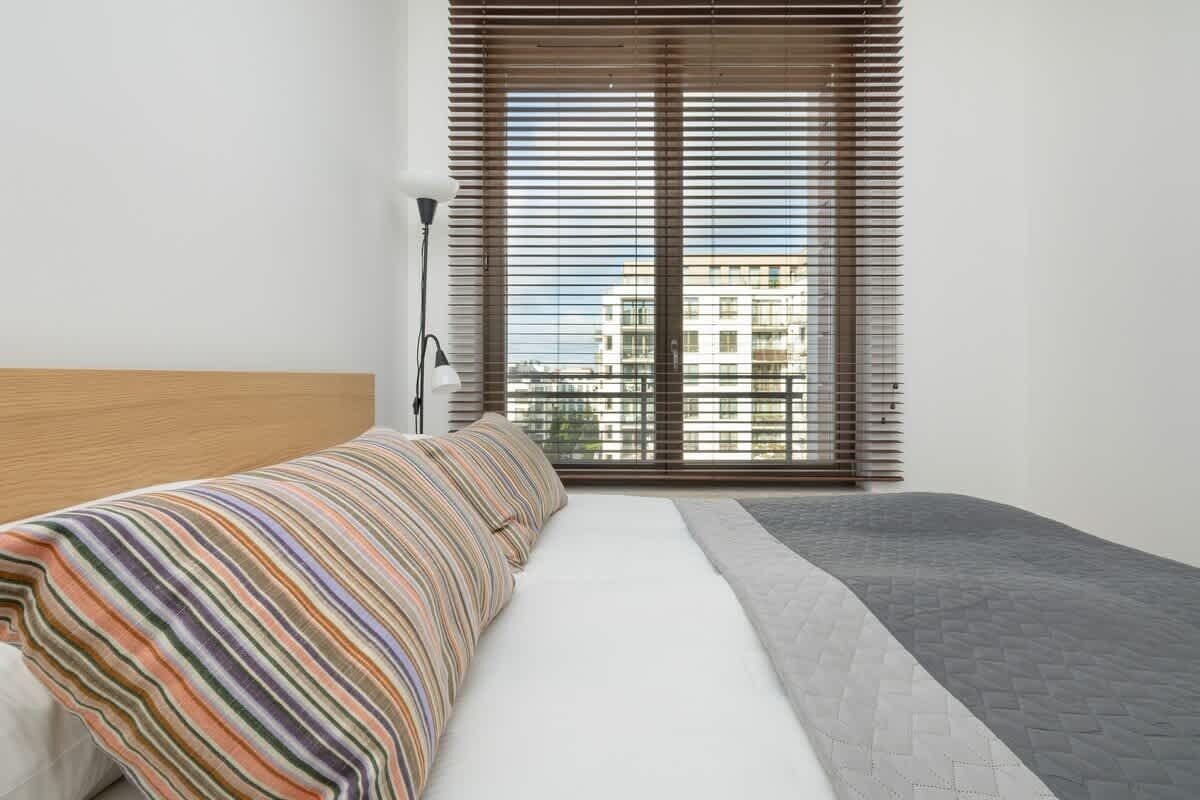 A modern bedroom with a striped bedspread, a wooden headboard, and a view of buildings through large windows with blinds.