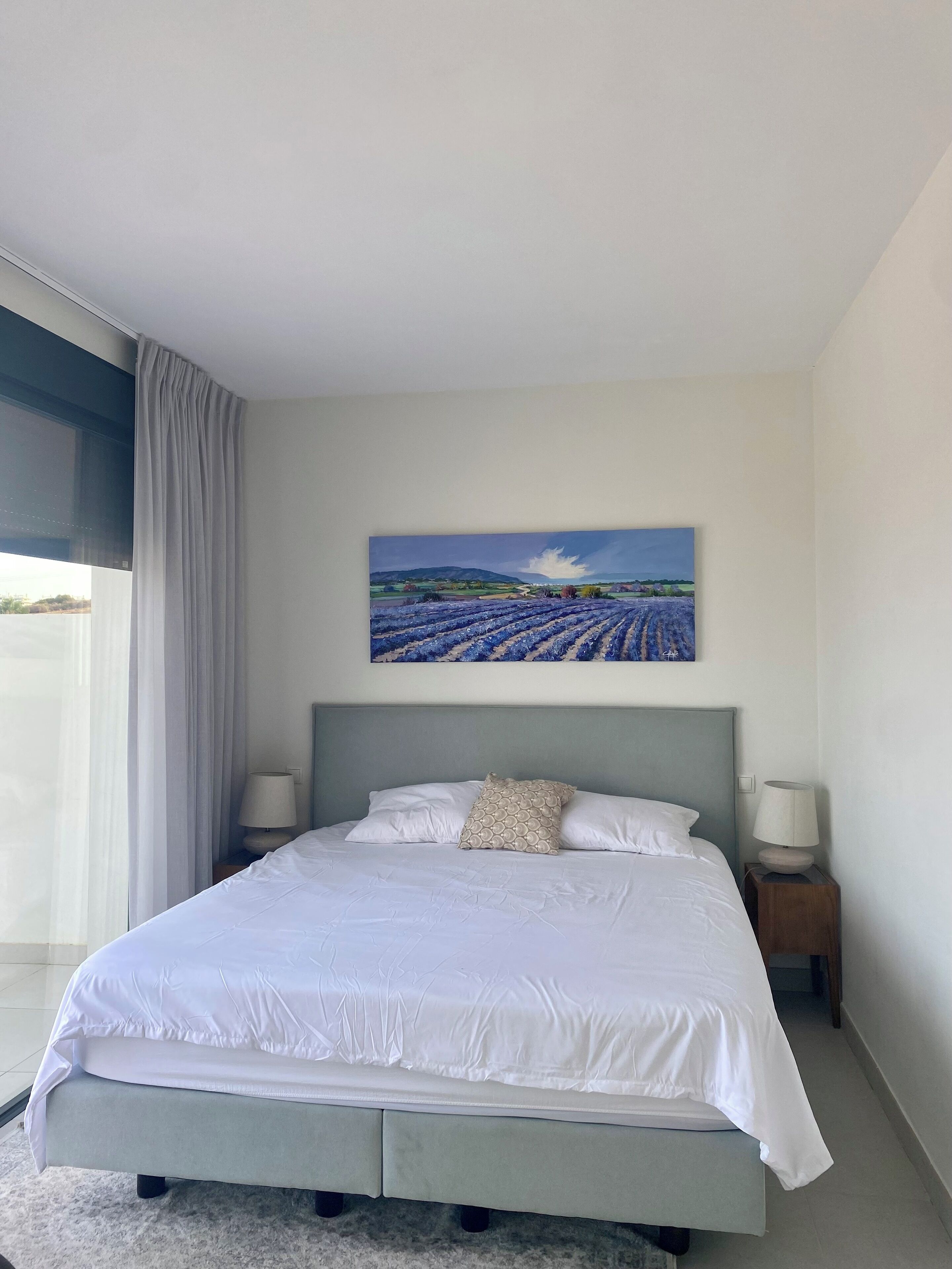 Une chambre moderne avec un grand lit, deux tables de chevet, un tableau représentant des champs de lavande et une vue dégagée par une grande fenêtre.