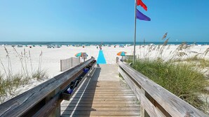 Une promenade en bois menant à une plage où l’on trouve des parasols et des personnes se prélassant au soleil.