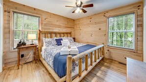 Dormitorio de cabaña de madera con cama, dos ventanas, una mesita de noche, ventilador de techo y vistas a árboles.