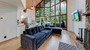 Un interior moderno con un gran techo de madera, un sofá modular azul oscuro, una chimenea de piedra y un amplio ventanal con vistas a la vegetación.
