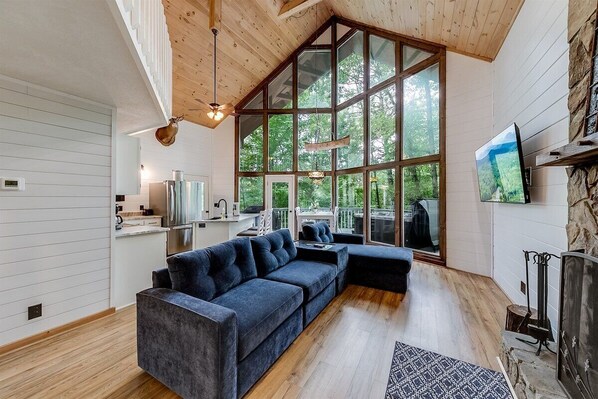 A modern interior with a large wooden ceiling, a dark blue sectional sofa, a stone fireplace, and a large window overlooking greenery. - Attitude Adjustment Mtn Chalet w/Private Hot Tub (Sautee Nacoochee)