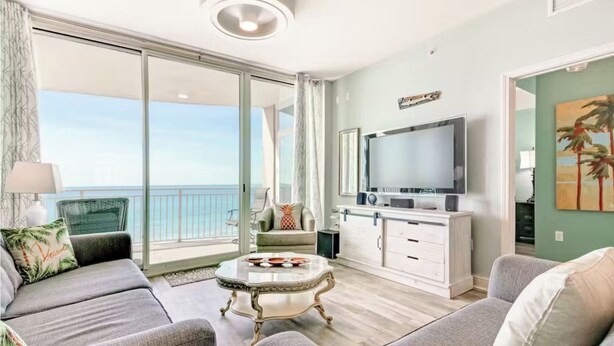 A living room with a sofa, a coffee table, a TV, and a balcony with a view of the ocean.