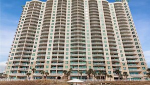 A high-rise hotel with multiple balconies, a beach view, and palm trees in the foreground.