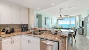 A modern kitchen with granite countertops, stainless steel appliances, and a dishwasher. There is a dining area with a table and chairs, and a view of the ocean through large windows.