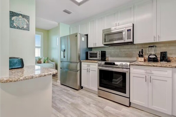 A modern kitchen with white cabinets, stainless steel appliances, a granite countertop, and a view of the living area.