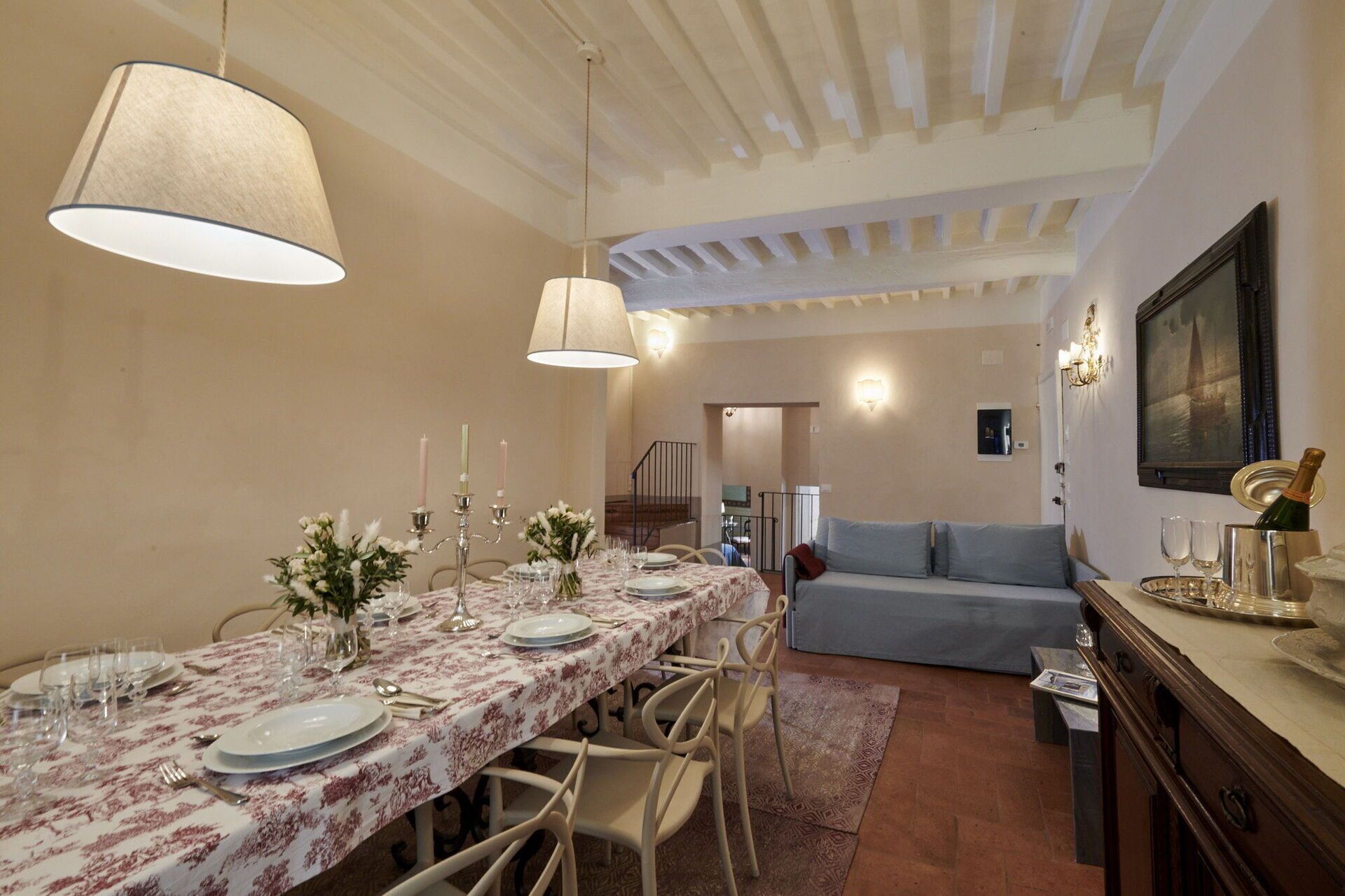 A dining area with a long table set for six, featuring a floral tablecloth, candles, and glassware. A sofa and a cabinet with a bottle of champagne are visible in the background.