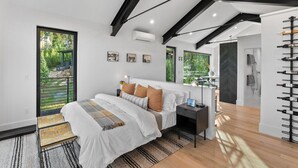 Un dormitorio moderno con una cama grande, una mesita de noche, una silla, una estantería para vinos y vistas a zonas verdes.