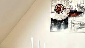 Ein Esstisch mit dunkler Holzoptik, weißen Stühlen und einer grauen Platzmatte. Drei weiße Kerzen stehen in schwarzen Kerzenhaltern, und ein Glasgefäß mit klarer Flüssigkeit steht daneben. Ein Metallschneebesen liegt auf der Platzmatte. An der Wand hinter dem Tisch hängt ein großes, abstraktes Gemälde.