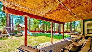 Ein überdachter Außenbereich mit Holzboden, Sitzgelegenheiten und Blick auf einen Garten mit einer Rutsche und Bäumen.