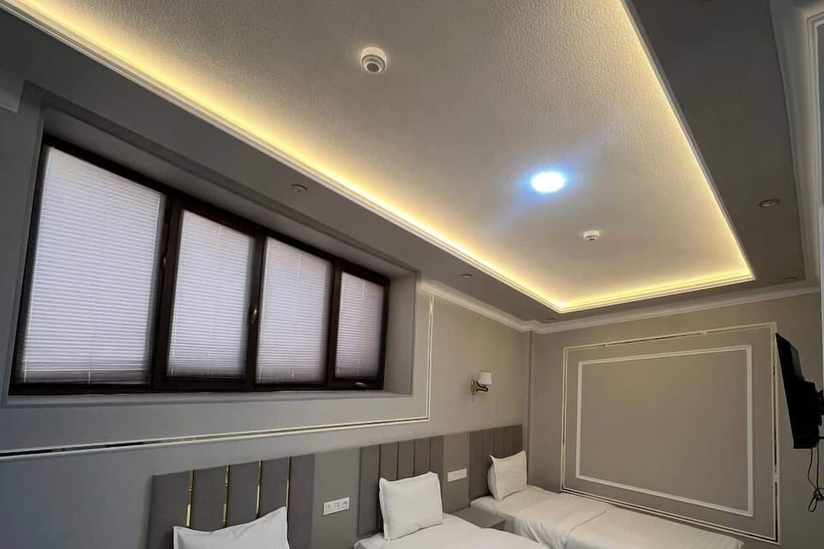 A hotel room with three single beds, a large window with blinds, a ceiling light, and a patterned carpet.