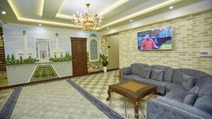 Un vaste espace de vie comprenant un grand canapé, une table basse en bois et un lustre. Un téléviseur est fixé au mur, et une fresque représentant une mosquée est visible.