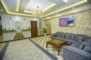 Un amplio salón con un sofá grande, una mesa de centro de madera y una araña. Un televisor está montado en la pared, y se observa un mural de una mezquita.