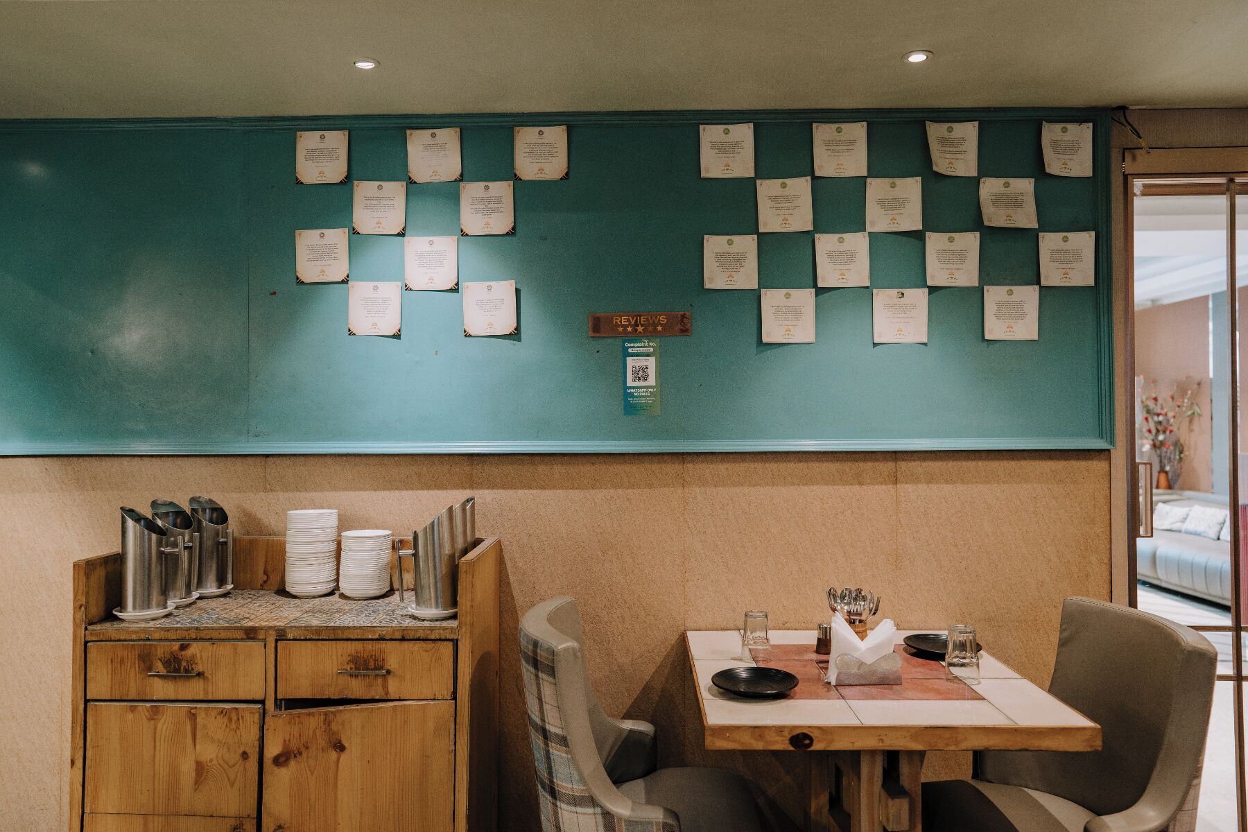 Interior de restaurante con una pared azul aguamarina mostrando numerosos certificados, un mueble de madera con utensilios de cocina y una mesa dispuesta para comer.