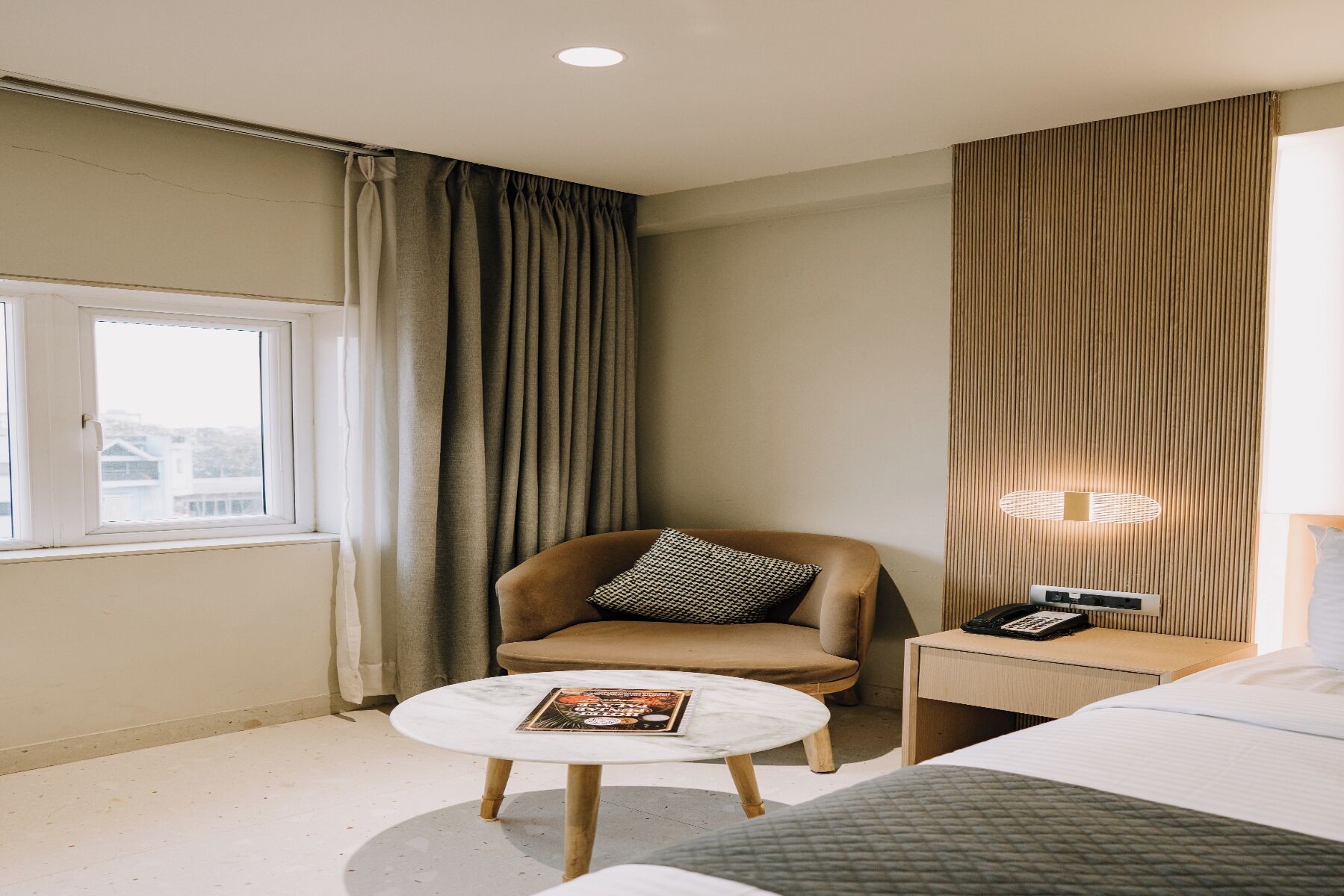 A hotel room with a bed, a round table with a magazine, a chair with a pillow, a bedside table with a phone, and curtains.