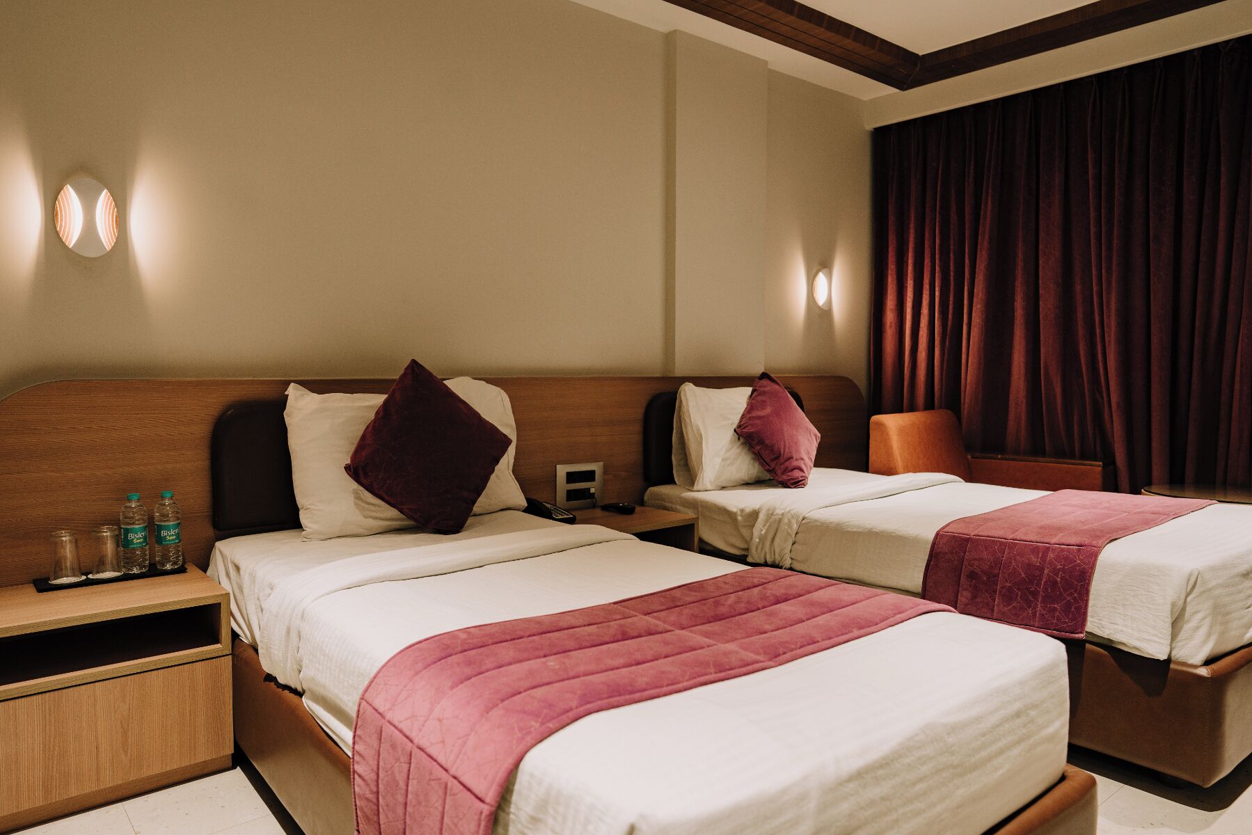 A hotel room with two beds, a wooden headboard, a nightstand with water bottles, and a curtain.