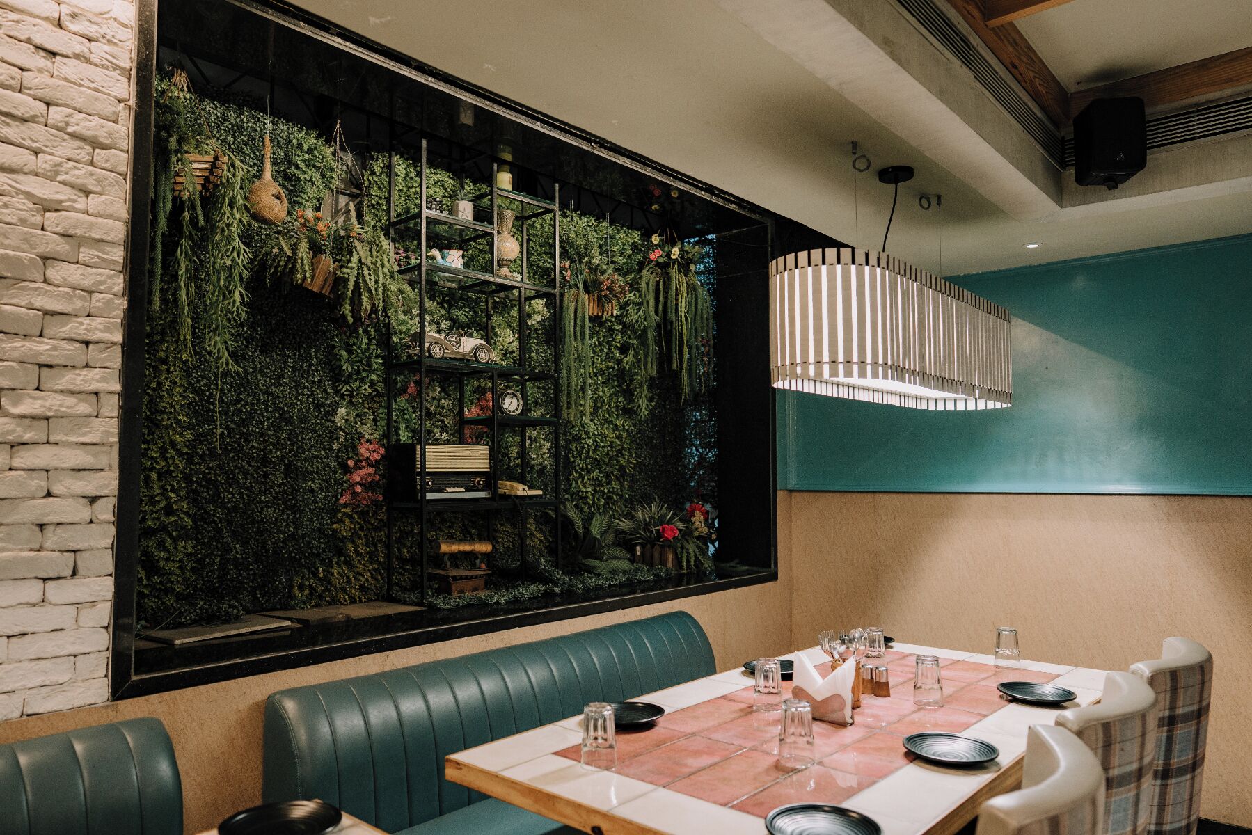 Un restaurante con asientos en una cabina verde, una mesa de madera dispuesta para comer y un amplio ventanal con pared verde y plantas colgantes.