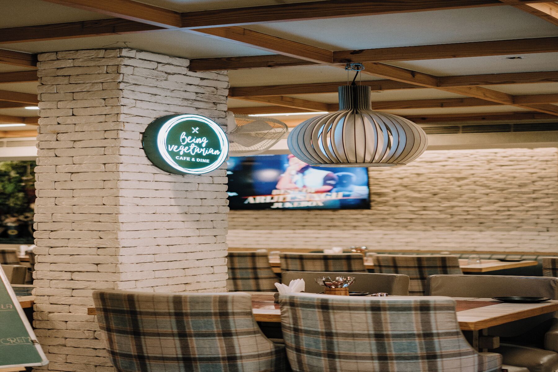 Interior de restaurante con un pilar de piedra, sillas a cuadros, una lámpara colgante y una pantalla mostrando un evento deportivo.