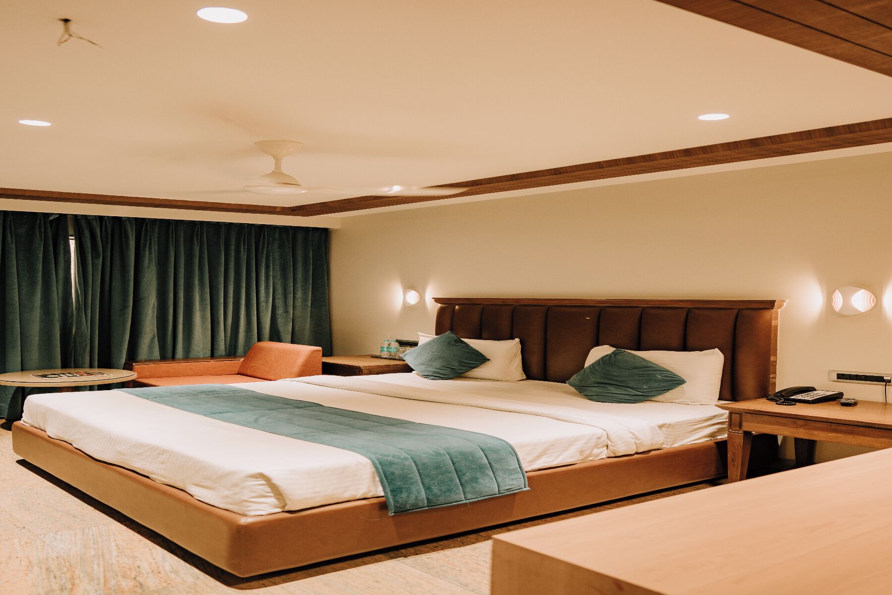 Una habitación de hotel con una cama grande, una mesita de noche, una silla, una pequeña mesa y un ventilador de techo.