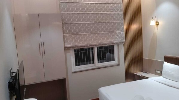 Una habitación de hotel con cama, armario, ventana con persianas, mesita de noche y lámpara de pared.