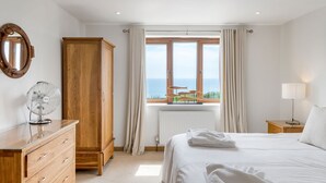 Un dormitorio con un armario de madera, una mesita de noche con lámpara, una cama con ropa blanca y vistas al mar a través de una ventana, además de un radiador.